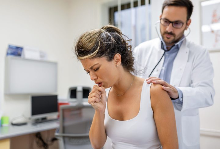 Young female patient in the clinic suffered from pneumonia, she is coughing the doctor listens to the wheezing in the lungs with a stethoscope.